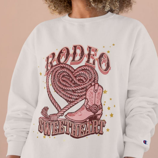 Woman wearing a blush sweatshirt with 'Rodeo Sweetheart' design on a beige background